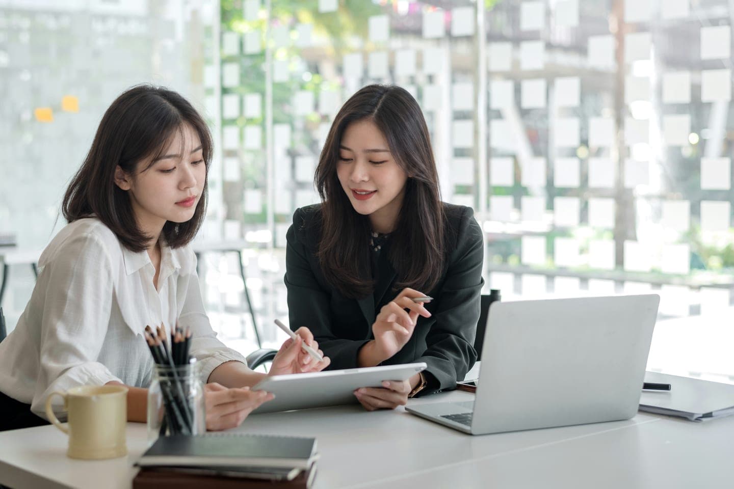 밝은 창가 책상에서 한 사람이 펜으로 태블릿을 가리키며 이야기하고 옆 사람이 받아 적는 상담 장면