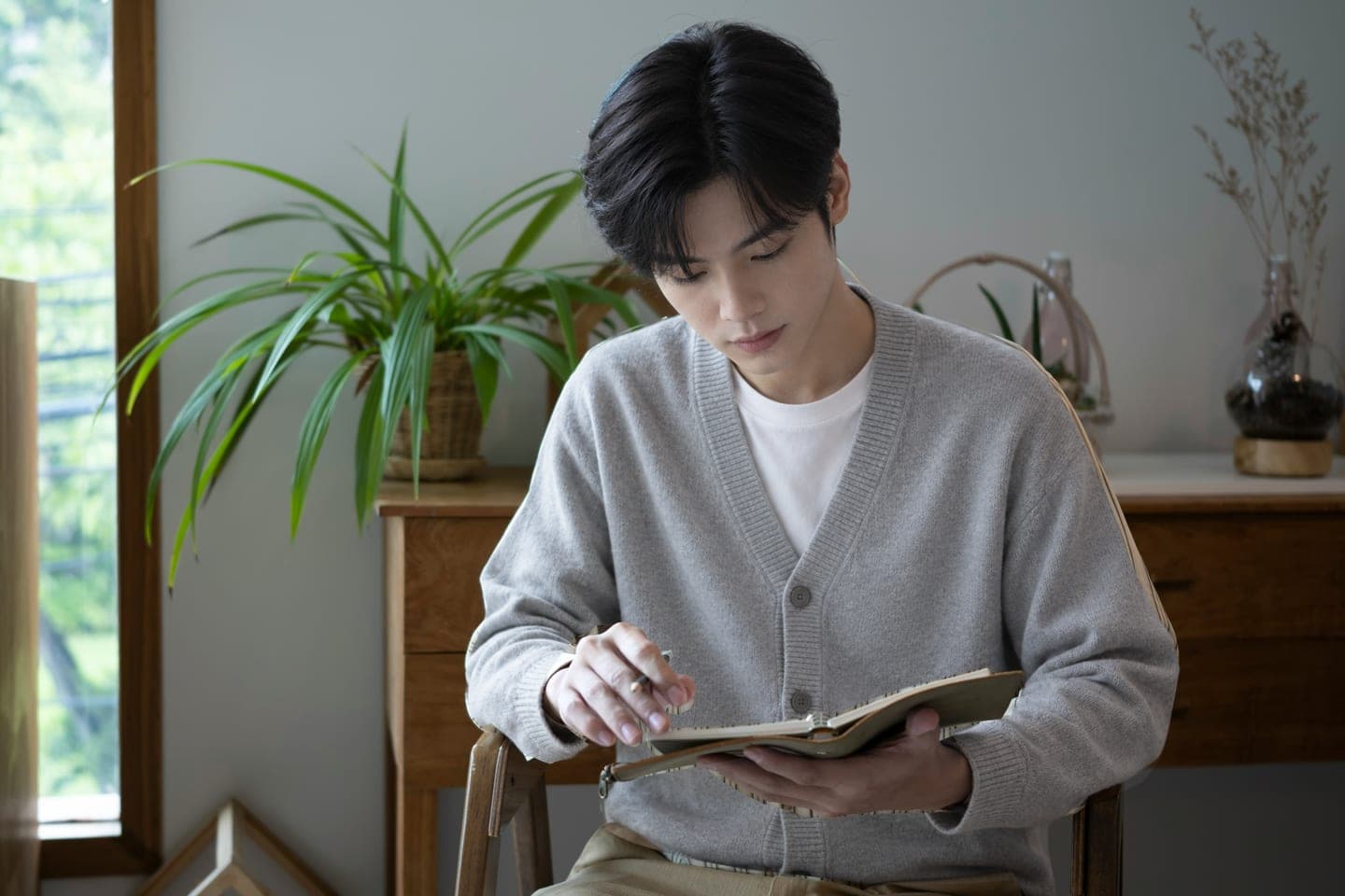 창가 화분 옆 의자에 앉아 펜을 쥔 채 펼친 수첩을 살펴보는 장면