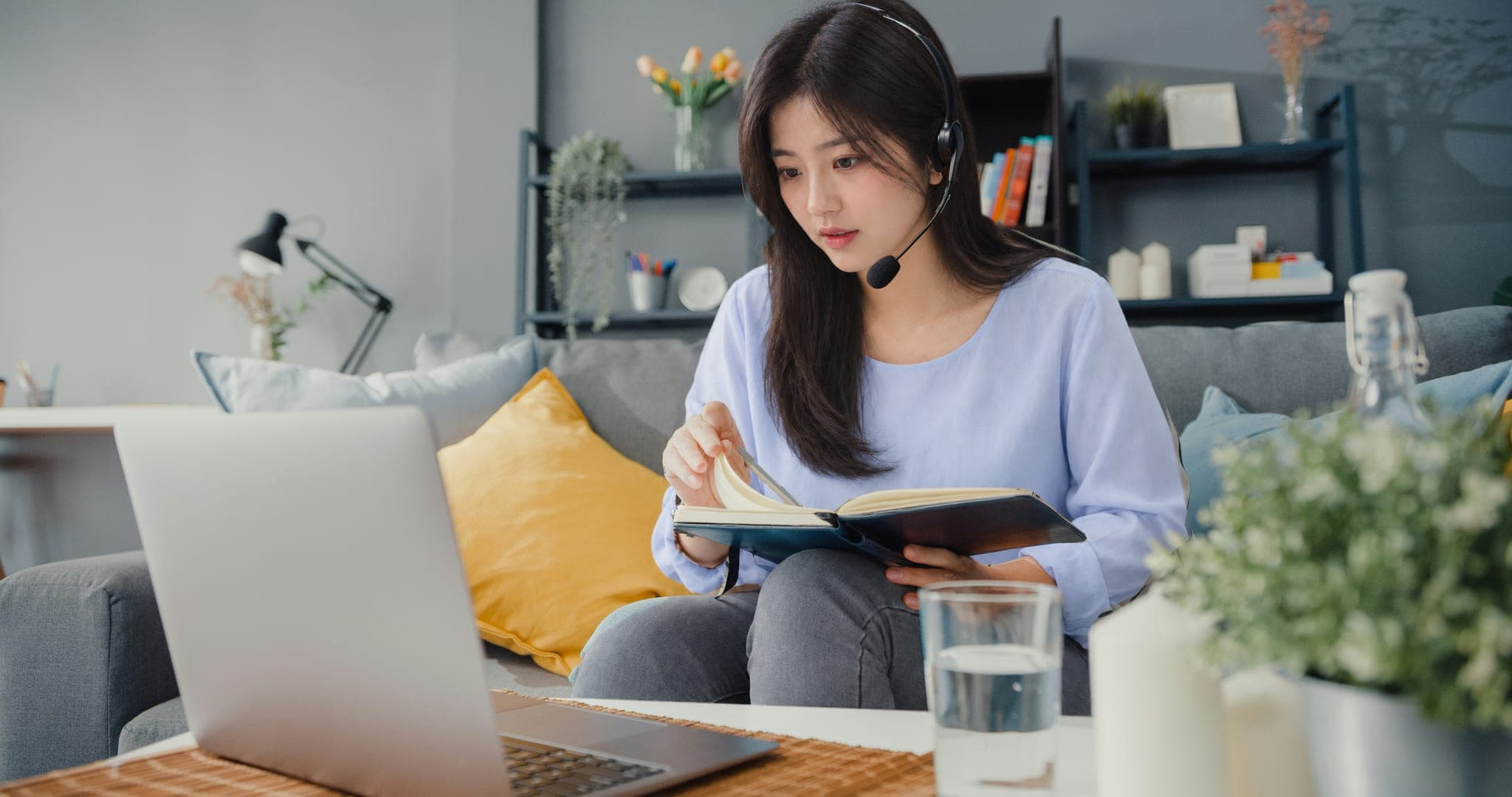 소파에 앉아 헤드셋을 쓰고 노트북 앞에서 펼친 책의 페이지를 넘기는 장면